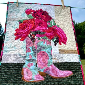 Puede incluir: Un tapiz de pared acolchado con un par de botas de vaquero con un estampado floral en tonos rosas y azules. Las botas parecen sostener un ramo de rosas rosas grandes y vibrantes. El fondo es un mosaico de colores y patrones.