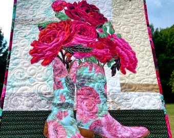 May include: A quilted wall hanging featuring a pair of light blue cowboy boots with a pink floral pattern, topped with a large bouquet of vibrant red and pink peonies. The background includes cream-colored fabric with swirling stitch patterns.