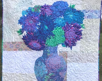 May include: A square quilt featuring a central floral arrangement of blue, purple, and green chrysanthemums in a matching patterned vase. The background consists of white fabric with a swirling quilted texture and horizontal bands of patterned fabric.