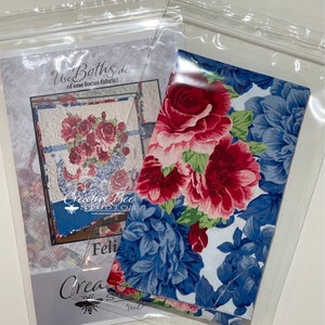 May include: A close-up of a fabric with a floral pattern. The fabric features large red and pink roses with green leaves on a white background. The fabric is folded in half and is in a clear plastic bag. The bag has a label that says "Use Both Sides of one focus fabric!" and "Creative Bee Studios".