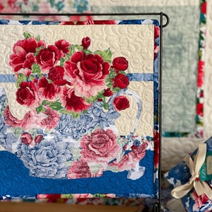 May include: A quilted wall hanging featuring a teapot filled with red, pink, and white roses. The teapot is blue and white with a floral pattern. The background is a cream color with a quilted pattern.