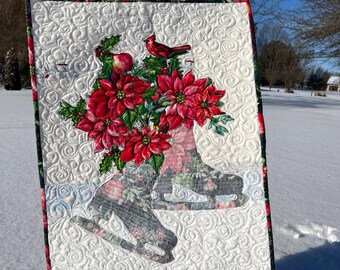 May include: A rectangular garden flag featuring a white quilted background with a swirling pattern. The design displays a pair of dark floral ice skates filled with vibrant red poinsettias, green holly leaves, and a small red cardinal bird perched on top.