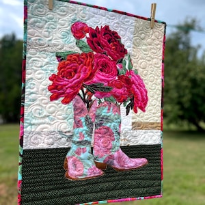 Könnte beinhalten: Ein gestepptes Wandbehang mit einem Paar Cowboy-Stiefeln, die mit leuchtend rosa Rosen gefüllt sind. Die Stiefel haben ein Blumenmuster in Rosa- und Türkistönen. Der Hintergrund ist cremefarben mit einem Steppmuster, und die Kanten sind mit Blumenstoff verziert.