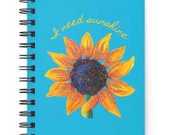 May include: A spiral-bound notebook with a bright blue cover featuring a painted sunflower with yellow and orange petals and a dark center. Above the flower, the text "I need sunshine" is written in a cursive yellow font. The notebook has a black wire coil.