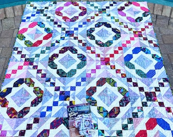 May include: A rectangular quilt featuring a repeating geometric pattern of light blue, pink, and multi-colored floral fabric blocks. A Quiltmaker magazine issue titled Bold & Beautiful is placed on the surface of the quilt near the bottom center.