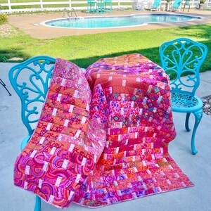 May include: A colorful quilt with a patchwork design, featuring shades of pink, orange, and red. The quilt is draped over a turquoise metal chair, with a swimming pool and green lawn in the background. The quilt's pattern includes geometric and floral elements.