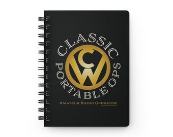 May include: A black spiral-bound notebook featuring a gold circular logo with the letters CW in the center. The text CLASSIC PORTABLE OPS is arched around the logo, with AMATEUR RADIO OPERATOR printed in gold below the central graphic design.