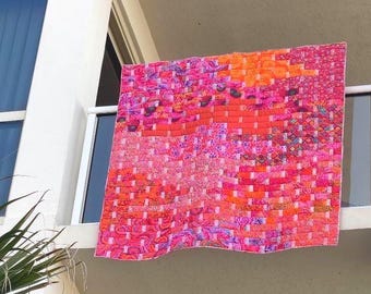 May include: A square quilt featuring a woven-style pattern constructed from various fabric strips in vibrant shades of hot pink, magenta, orange, and coral. The textile displays a complex grid of interlocking colors and diverse floral patterns.