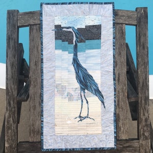 May include: A blue heron quilt block with a white background and a blue border. The heron is standing on a beach with seashells and the text "Creative Bee Quilts" is in the lower right corner.