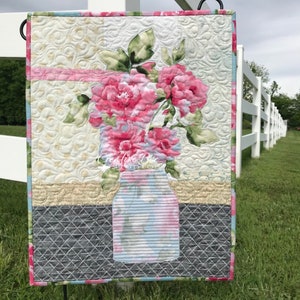 Może przedstawiać: Flaga ogrodowa pikowana z różowym wzorem kwiatowym w niebieskim i białym słoiku. Flaga jest zawieszona na białym drewnianym płocie z zieloną trawą na pierwszym planie.