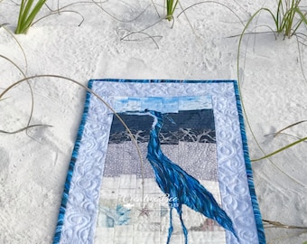 May include: A rectangular fabric wall hanging featuring a blue heron appliqué centered on a pieced background of light blue, gray, and white squares. The border is quilted with a swirling pattern and the text Creative Bee Studios is visible.