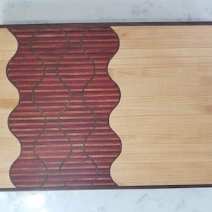 Puede incluir: Una tabla de cortar de madera con un diseño ondulado. El lado izquierdo de la tabla está hecho de madera oscura con un patrón de rayas, mientras que el lado derecho está hecho de madera clara.