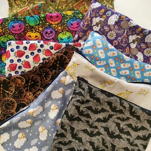 May include: A selection of zipped pouches featuring diverse, colourful patterns. Designs include pumpkins, strawberries, clocks, sheep, bats, and other whimsical motifs. The pouches are made of fabric and are rectangular.