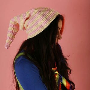 May include: A handmade, long, striped hat in soft pastel colours of pink and yellow. The hat is crocheted and has a long, pointed end. The hat is worn by a person with long, dark hair. The background is a solid pink.