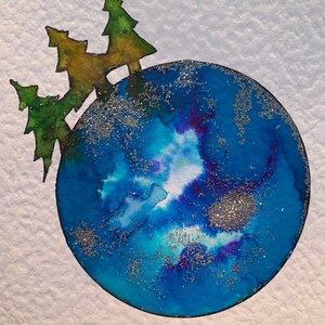 May include: A blue watercolor painting of a globe with silver glitter. Three green and brown trees are drawn on top of the globe.