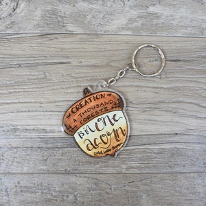 May include: A brown and tan acorn-shaped keychain with a silver chain. The keychain features the quote "The creation of a thousand forests is in one acorn." by Ralph Waldo Emerson.