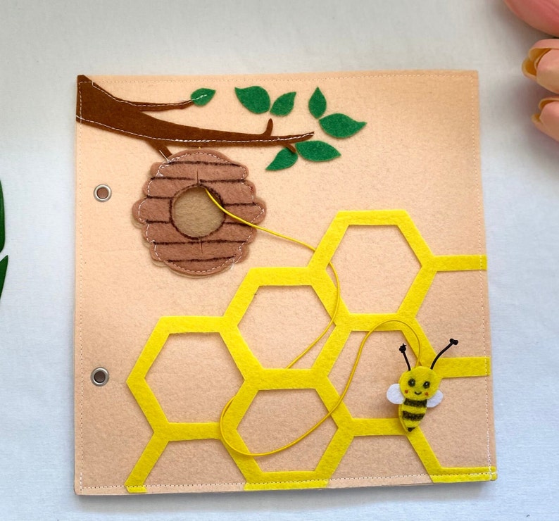May include: A felt beehive activity board with a yellow honeycomb, a brown felt beehive, and a yellow felt bee with a black and white striped body. A string is attached to the beehive and can be threaded through the honeycomb.