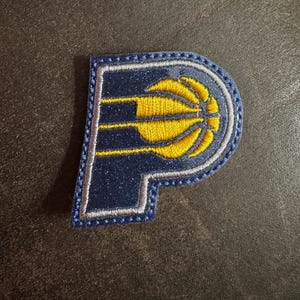 May include: A navy blue embroidered patch in the shape of the letter "P". The patch features a yellow basketball design with three horizontal lines. The edges are outlined in silver and blue stitching.