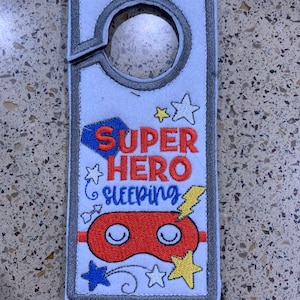May include: A white door hanger with a gray border. The hanger has a hole at the top for hanging on a door. The hanger has an embroidered design of a red sleep mask with white eyes, stars, and the text "SUPER HERO SLEEPING".