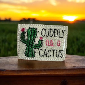 May include: A white, glittery patch with a green cactus and pink flowers. The text "CUDDLY as a CACTUS" is embroidered in black thread.