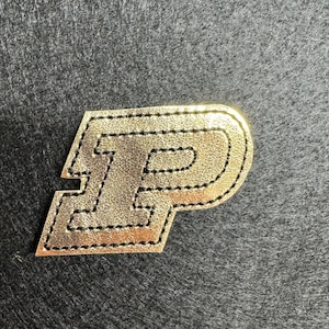 May include: A gold-colored leather patch with a black stitched outline in the shape of the letter "P".