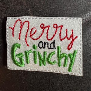 May include: A rectangular white patch with the words "Merry and Grinchy" embroidered in red, green, and black thread. The patch has a stitched border and is set against a dark background.