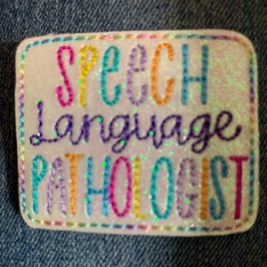 May include: A rectangular, glittery badge with the words "SPEECH LANGUAGE PATHOLOGIST" embroidered in various colors. The badge has a white background and a colorful stitched border.