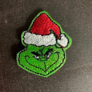 May include: Green felt Grinch face with a red and white Santa hat. The Grinch is smiling with a mischievous look.