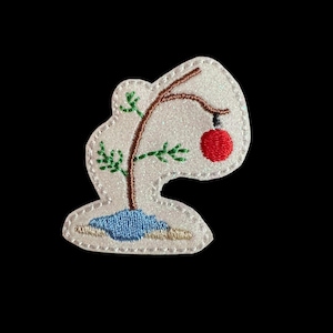 May include: An embroidered patch featuring a whimsical Christmas tree design. The tree has a brown trunk and branches with green leaves, a red ornament, and a blue base. The patch has a white, glittery background.