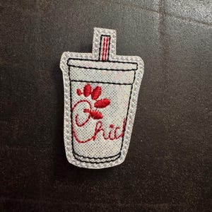May include: A white glittery embroidered patch in the shape of a beverage cup. The cup has a red paw print and the word "Chick" in red script. The cup has a black outline and a red and white straw.