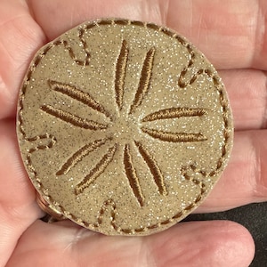 May include: A beige sand dollar with a brown embroidered design. The sand dollar is glittery and has a stitched edge.
