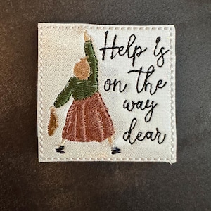 May include: Embroidered patch with a woman in a green top and a rust-colored skirt, reaching upwards. The text "Help is on the way dear" is written in black script on a white background.