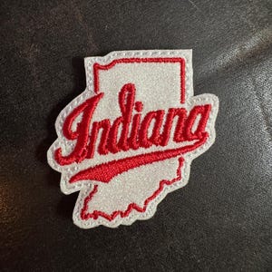 May include: A white and red embroidered patch in the shape of the state of Indiana. The word "Indiana" is written in red cursive script across the center of the patch. The patch has a white border.