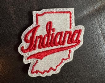 May include: A white and red embroidered patch in the shape of the state of Indiana. The word "Indiana" is written in red cursive script across the center of the patch. The patch has a white border.