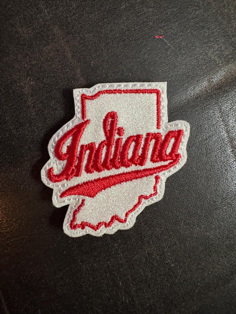 May include: A white and red embroidered patch in the shape of the state of Indiana. The word "Indiana" is written in red cursive script across the center of the patch. The patch has a white border.