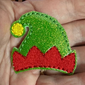 May include: A green glittery elf hat with a red zig-zag trim and a yellow pom-pom. The hat is stitched with a contrasting thread. This festive accessory is perfect for holiday crafts and costumes.