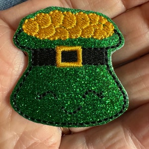 May include: A green felt leprechaun hat with gold coins and a black belt. The hat is decorated with glitter.