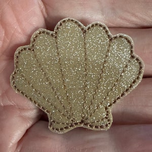 May include: A gold glitter seashell with a brown stitched outline. The seashell is a decorative embellishment for crafts and projects.