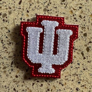 May include: A red and white embroidered patch featuring the Indiana University logo. The logo is a stylized letter "I" with a vertical line through the center and two vertical lines on either side. The patch has a red border and white stitching.