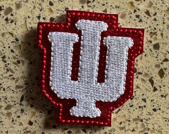 以下が含まれることがあります： インディアナ大学のロゴが入った赤と白の刺繍パッチ。ロゴは、中央に縦線、両側に2本の縦線が入った、様式化された「I」です。パッチは赤い縁取りと白いステッチが施されています。