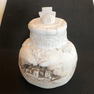 May include: White ceramic jar with a lid featuring a stylized design. The jar has a textured surface and a raised design around the top.