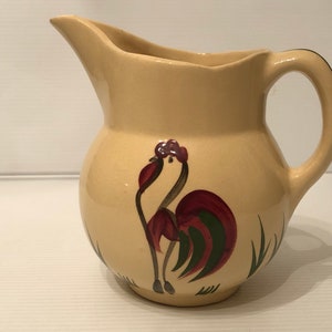 May include: A cream-colored ceramic pitcher with a rooster design. The rooster is painted in shades of red, green, and brown. The pitcher has a rounded body and a handle.