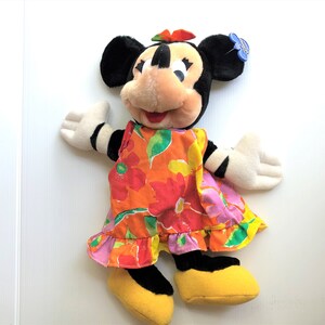 以下が含まれることがあります： カラフルな花柄のドレスを着たミニーマウスのぬいぐるみ。耳は黒く、赤いリボン、白い顔に黒い目と赤い口をしています。