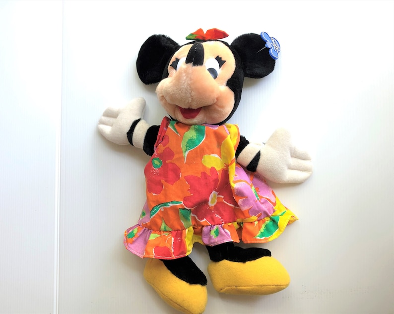 以下が含まれることがあります： カラフルな花柄のドレスを着たミニーマウスのぬいぐるみ。耳は黒く、赤いリボン、白い顔に黒い目と赤い口をしています。
