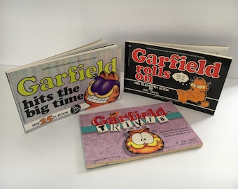 May include: Three vintage Garfield books are displayed. The titles include "Garfield hits the big time", "Garfield rolls on", and "The Garfield Trivia". The books feature the cartoon cat Garfield.