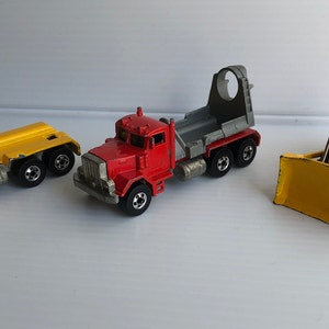 May include: Three toy construction vehicles: a yellow dump truck, a red semi-truck with a gray trailer, and a yellow bulldozer.