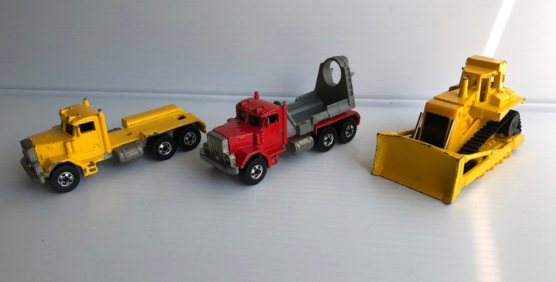 May include: Three toy construction vehicles: a yellow dump truck, a red semi-truck with a gray trailer, and a yellow bulldozer.