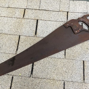 May include: A rusty, antique handsaw with a wooden handle. The handle has a carved design and a metal ring. The saw is lying on a gray surface.
