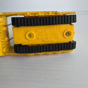 May include: Yellow toy bulldozer with black tracks. The bottom of the toy has the text "HOT WHEELS&reg;  CAR BULLDOZER &copy;MATTEL INC. 1979 MADE IN TAIWAN".