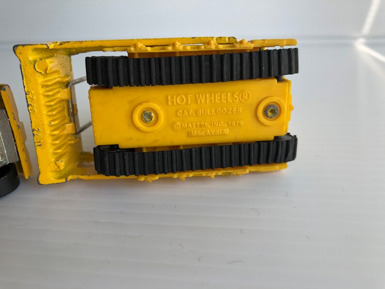 May include: Yellow toy bulldozer with black tracks. The bottom of the toy has the text "HOT WHEELS&reg;  CAR BULLDOZER &copy;MATTEL INC. 1979 MADE IN TAIWAN".